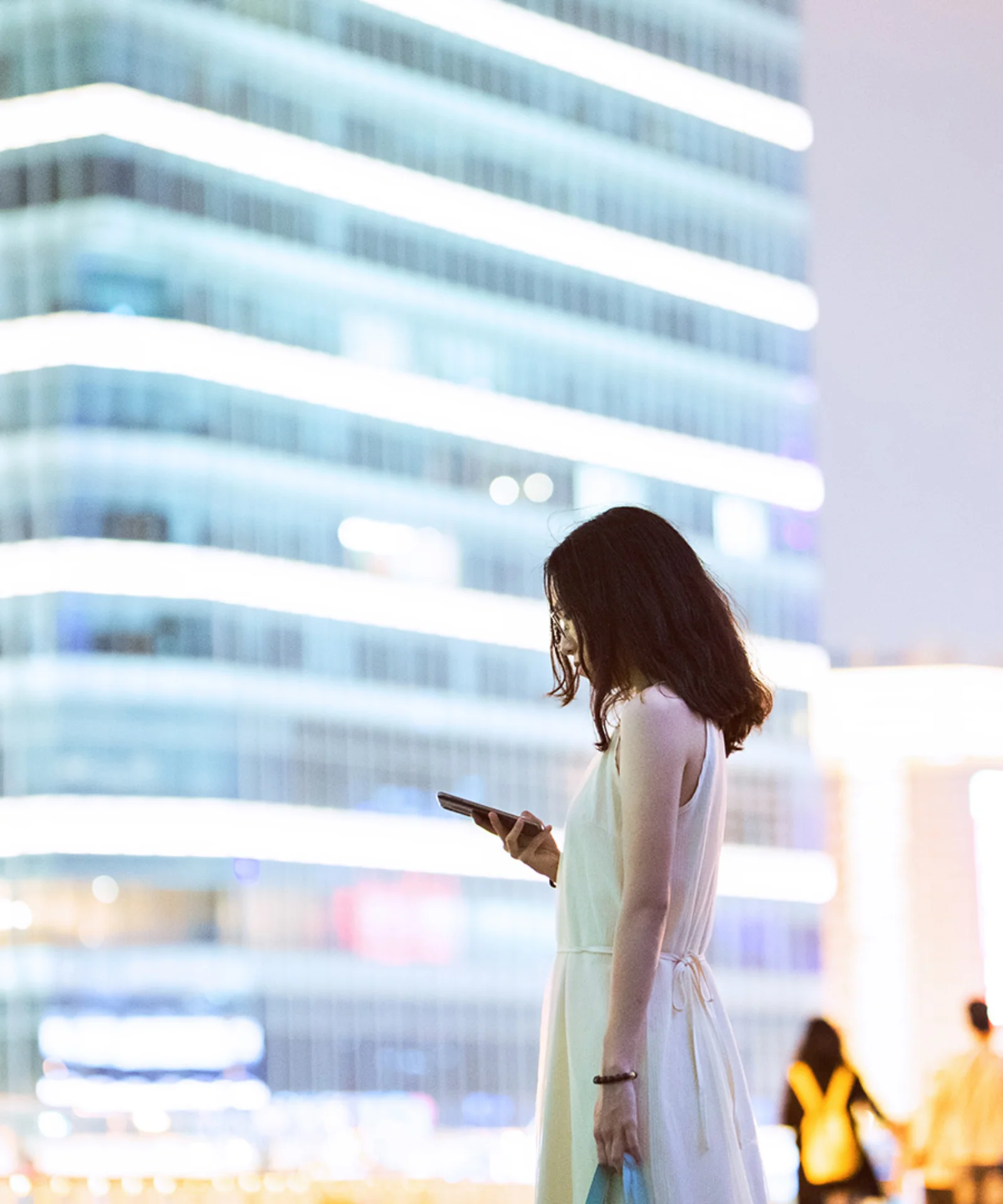 Uma mulher parada em um ambiente urbano à noite, olhando para seu smartphone, com prédios modernos bem iluminados ao fundo, representando o tema da digitalização no setor bancário.