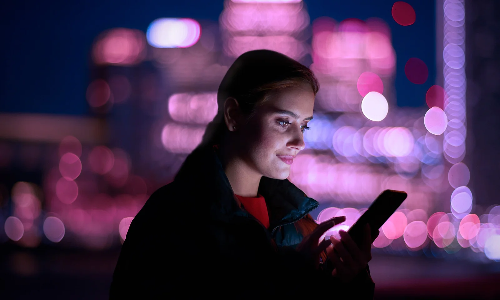 Taking Guidewire to the Cloud A woman using a smartphone against a backdrop of a cityscape illuminated by pink and purple lights, symbolising the transition of Guidewire to cloud-based solutions.