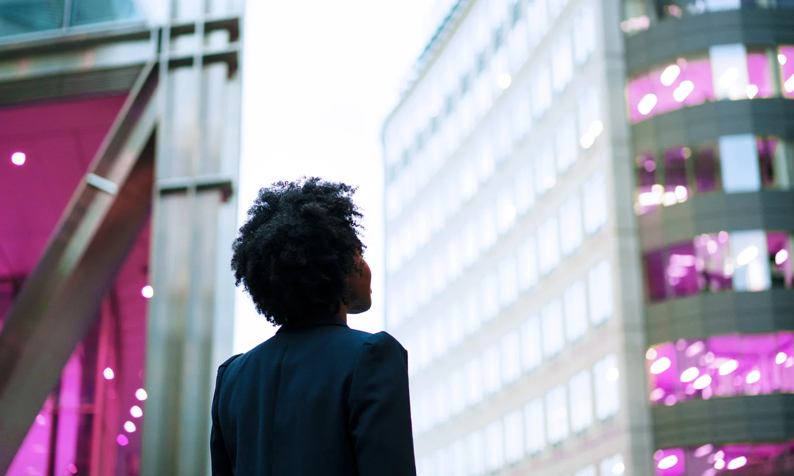 Visionary Leadership in the Modern Business Landscape A person with natural hair, wearing a dark blazer, gazing up at modern office buildings with illuminated windows in a city environment.